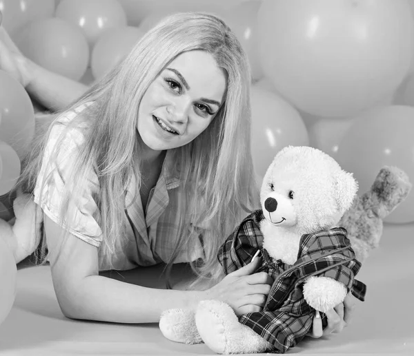 Blonde on smiling face relaxing with teddy bear toy. Woman cute celebrate birthday with balloons. Girl in pajama, domestic clothes lay near air balloons, pink background. Birthday girl concept