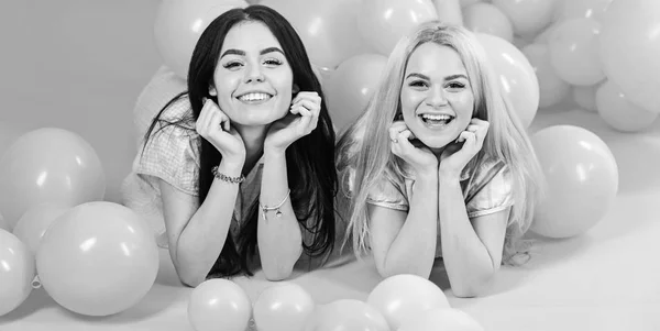 Les filles s'allongent sur le ventre près des ballons, fond rose. Soeurs ou meilleurs amis en pyjama à la soirée pyjama fille. Concept de commérage. Blonde et brune sur des visages souriants s'amusent à la fête de chambre domestique — Photo