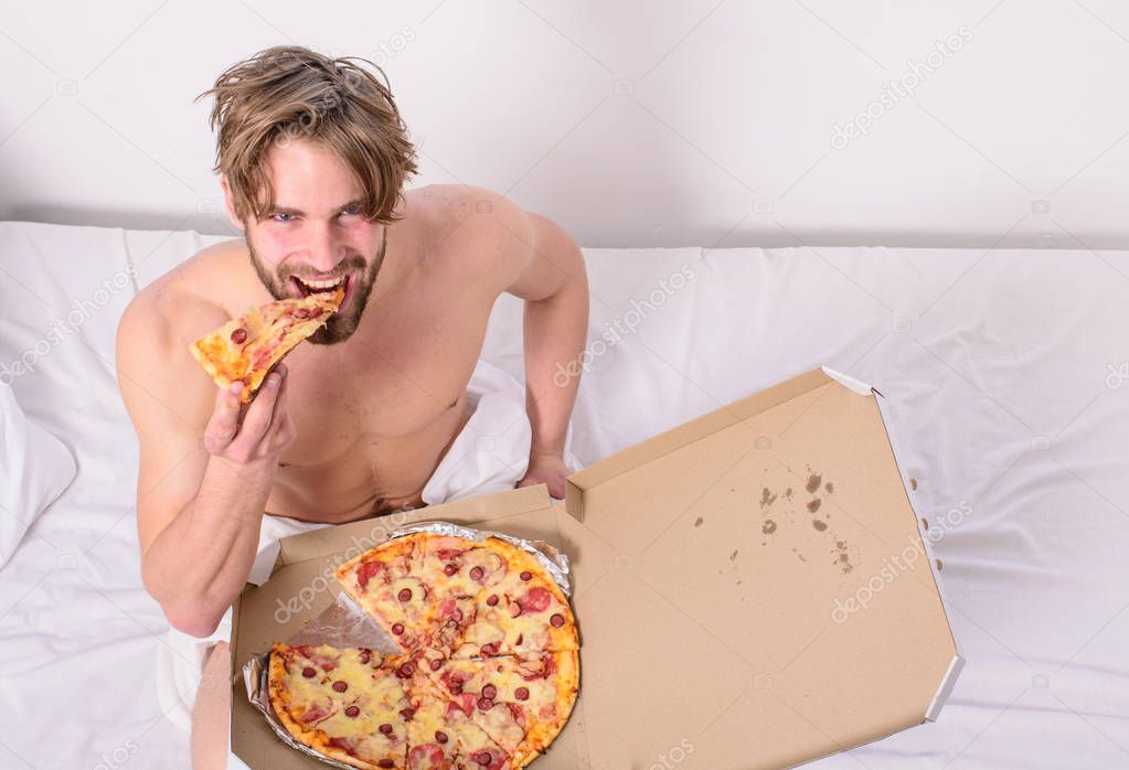 Man likes pizza for breakfast. Break diet concept. Guy holds pizza box sit bed in bedroom or hotel room. Food delivery service. Man bearded handsome bachelor eating cheesy food for breakfast in bed
