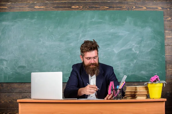 Revisa los deberes. El papeleo forma parte de la vida de los profesores. Profesora de escuela revisando tarea o prueba. Profesor sentarse escritorio con portátil. Maestro barbudo hipster con anteojos sentarse en el aula pizarra fondo — Foto de Stock