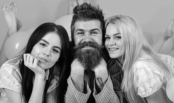 L'uomo con la barba e i baffi attrae ragazze bionde e brune. Le ragazze si innamorano di macho barbuto, sfondo rosa. Concetto maschio alfa. Trio su facce sorridenti giaceva vicino palloncini — Foto Stock