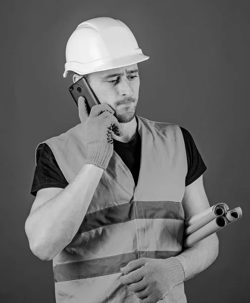 Engineer, architect, builder on busy face speaks on smartphone while holds blueprints. Architect concept. Man, foreman in helmet supervises construction on phone, red background