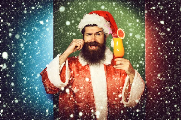 Knappe bebaarde kerstman met lange baard op glimlachend gelaat met glas alcoholvrije cocktail in kerst- of kersttrui en nieuwjaarshoed op kleurrijke studioachtergrond — Stockfoto