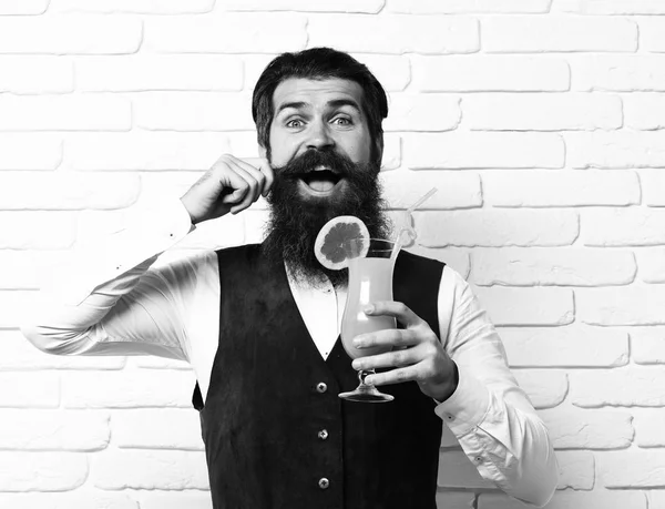 handsome bearded man with long beard and mustache has stylish hair on happy face holding glass of alcoholic beverage in vintage suede leather waistcoat on white brick wall studio background
