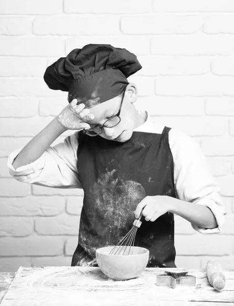 Kleiner Junge netter Koch Koch in roter Uniform und Hut auf gefärbtem Gesicht Mehl mit Gläsern am Tisch stehend mit Nudelholz und Kochen mit Küchenbesen in türkisfarbener Schüssel auf weißem Backsteinmauerhintergrund — Stockfoto