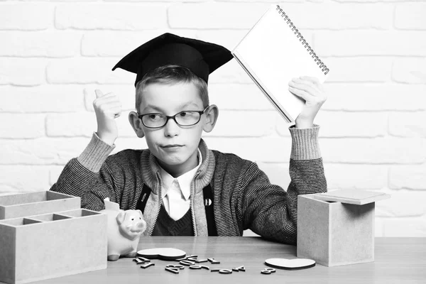 Ung söt elev pojke i grå tröja och glasögon sitter vid skrivbordet med copybook i handen trä siffror rosa gris bank i examen mössa i klassrummet visar cool på vit tegelvägg bakgrund — Stockfoto
