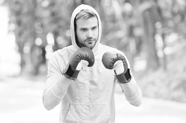 Training for big day. Man athlete on concentrated face with sport gloves practicing boxing punch, nature background. Boxer hood head practices jab punch. Sportsman boxer training with boxing gloves