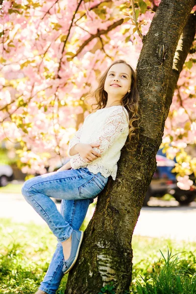 Dziewczyna na uśmiechniętą twarz, stojący w pobliżu sakura, oprzeć się na pniu drzewa. Dziewczyna z długimi włosami odkryty, Wiśniowy kwiat lub sakura kwiatami na tle. Koncepcja mood wiosna. Słodkie dzieci cieszyć się przyrodą na dzień wiosny — Zdjęcie stockowe