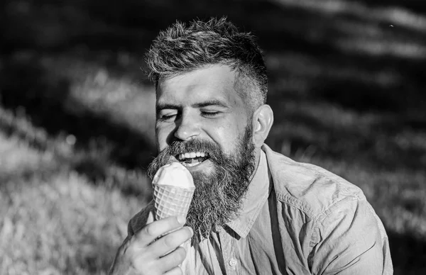Conceito de tentação. Homem barbudo com cone de gelado. Homem com barba e bigode no rosto feliz desfrutar de sorvete, grama no fundo, desfocado. Homem com barba longa come sorvete, enquanto se senta na grama — Fotografia de Stock