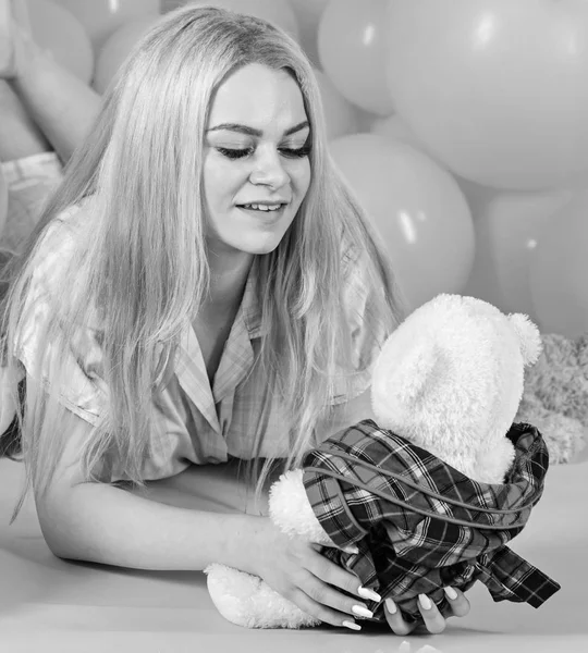 Блондинка на усміхненому обличчі розслабляється іграшкою плюшевого ведмедя. Дівчина в піжамі, домашній одяг лежала біля повітряних кульок, рожевий фон. Жінка мило відзначає день народження повітряними кулями. Концепція дівчини на день народження — стокове фото