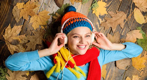 Girl cheerful face lay on wooden background with leaves top view. Autumn melody concept. Hipster woman knitted hat and scarf listen music headphones. Fall cozy atmosphere. Fall and autumn season Stock Photo