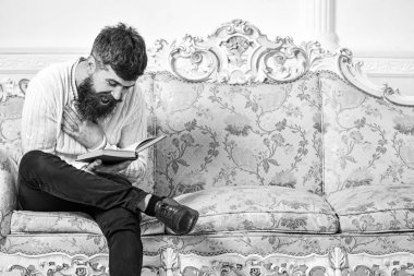 Adam zevk ile eski kitap okuma. Mizahi edebiyat kavramı. Maço üzerinde yüz kitap okuma gülüyor. Barok tarzı kanepe, tutar kitap, beyaz duvar arka plan sakal ve bıyık oturur adam