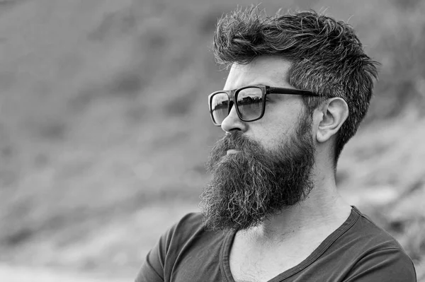 Hipster man in trendy sunglasses standing on slope with lush green glass in spring. Bearded man with stylish hairstyle enjoying the view over city