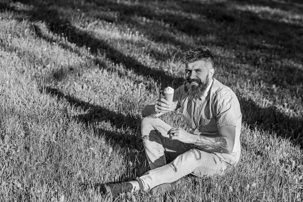 Man with long beard enjoy ice cream, while sits on grass. Delicacy concept. Man with beard and mustache on happy face eats ice cream, grass on background, defocused. Bearded man with ice cream cone