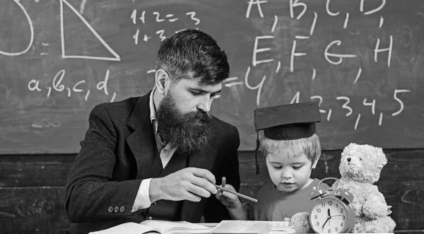Mathematics lesson concept. Father teaches son mathematics. Teacher in formal wear and pupil in mortarboard in classroom, chalkboard on background. Enthusiastic kid studying with teacher