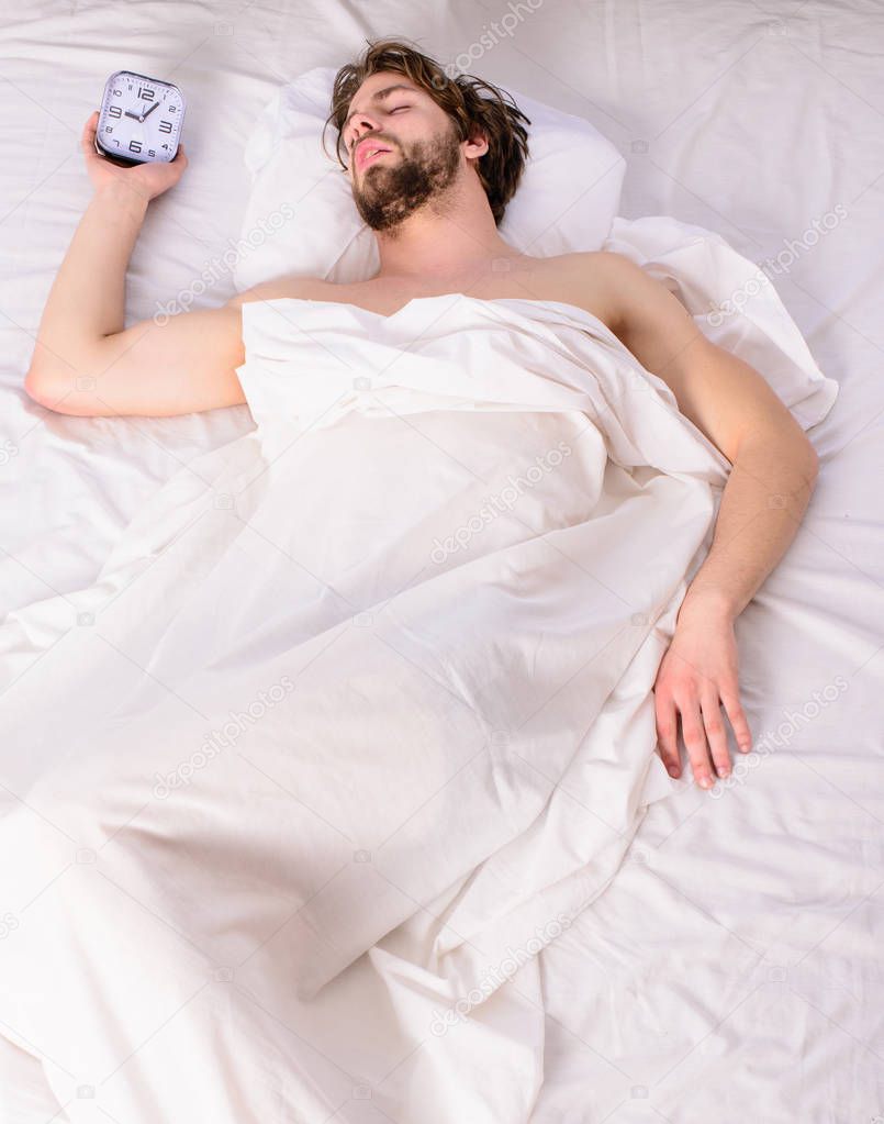 Fresh bedclothes concept. Man sleepy drowsy unshaven bearded face covered with blanket having rest. Turn off alarm clock. Man unshaven relaxing bed hold alarm clock. Guy lay under white bedclothes