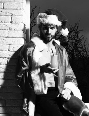 Genç, yakışıklı, sakallı, seksi Noel Baba sakallı, kırmızı Noel Baba şapkalı ve ceketli, kaslı, çıplak gövdeli, siyah ve tuğla duvar arkasında alkollü turuncu bir içecek bardağı tutan adam.
