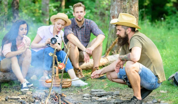 Grupo de amigos turista relaxante perto de fogueira. Homem assar salsicha enquanto os amigos falam compartilhar impressão e assistir câmera de fotos. Turistas que têm tempo de lanche com assado sobre comida de fogo. Caminhada florestal — Fotografia de Stock