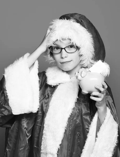 Joven lindo santa claus chico con gafas en suéter rojo y año nuevo sombrero de Navidad celebración de la hucha de cerdo rosa con la mano levantada sobre fondo de estudio azul —  Fotos de Stock