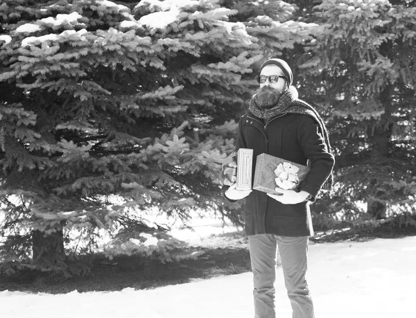 Hombre barbudo guapo, hipster, con barba y bigote en gafas de sol negras, sombrero, abrigo y bufanda con cajas de regalo como regalos en el día de invierno al aire libre sobre fondo natural —  Fotos de Stock