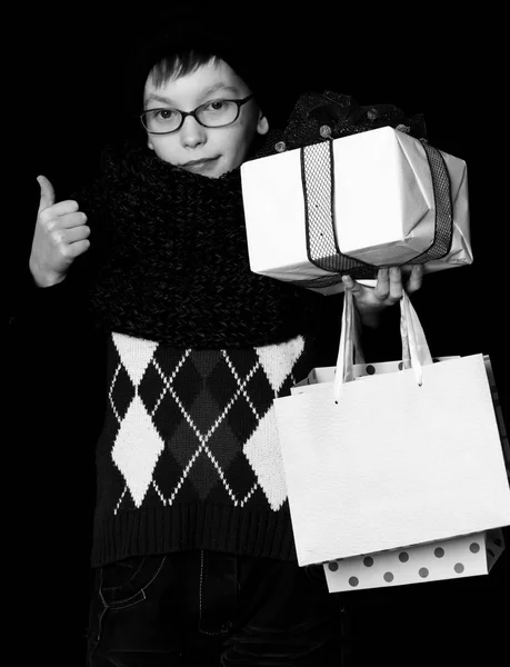 Pequeño niño sonriente o lindo niño nerd en gafas, sombrero y bufanda de punto de moda sobre fondo negro sostiene caja de regalo blanco con lazo rojo y bolsas de compras, sostiene el pulgar u — Foto de Stock