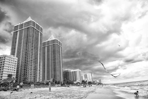 Miami, EE.UU. - 10 de enero de 2016: playa de mar y cometa vuelan en el cielo nublado. Edificios de apartamentos en la costa tropical. Arquitectura y bienes raíces en resort de lujo. Vacaciones de verano en el mar. Wanderlust y viajes — Foto de Stock