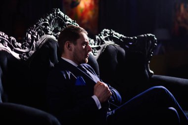 Patron şık elbiseli kendinden emin görünüyor. Zengin yaşam tarzı, profesyonel başarı, şık mobilyalar, para, zarafet kavramı. Adam ciddi bir yüz. İşadamı zarif vintage koltukta oturur ve düşünüyor