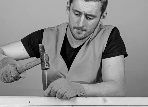 Carpenter, woodworker on concentrated face hammering nail into wooden board. Man, handyman in bright vest and protective gloves handcrafting, light blue background. Handcrafting concept — Stock Photo, Image