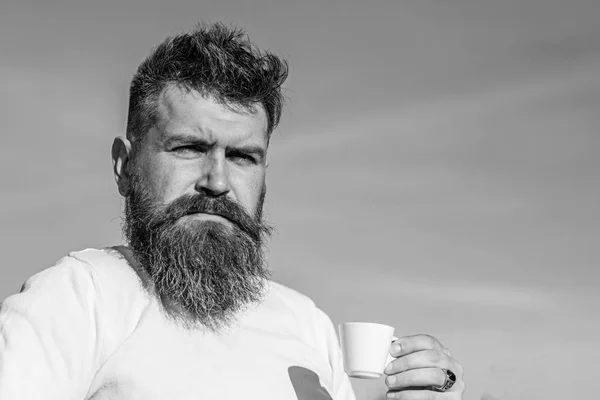 Man with long beard enjoy coffee. Coffee gourmet concept. Bearded man with espresso mug, drinks coffee. Man with beard and mustache on strict face drinks coffee, blue sky background, defocused