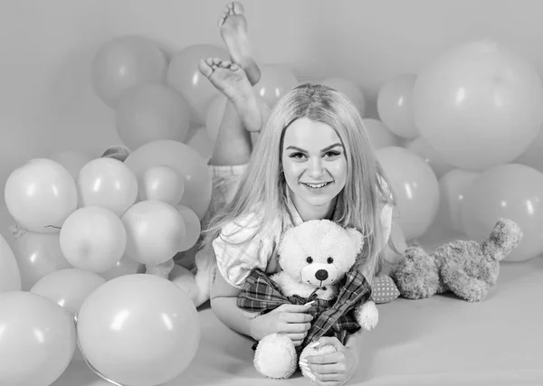 Woman cute celebrate birthday with balloons. Girl in pajama, domestic clothes lay near air balloons, pink background. Birthday girl concept. Blonde on smiling face relaxing with teddy bear toy