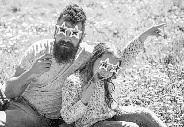 Rock star concept. Family spend leisure outdoors. Father and daughter sits on grass at grassplot, green background. Child and dad posing with star shaped eyeglases photo booth attribute at meadow