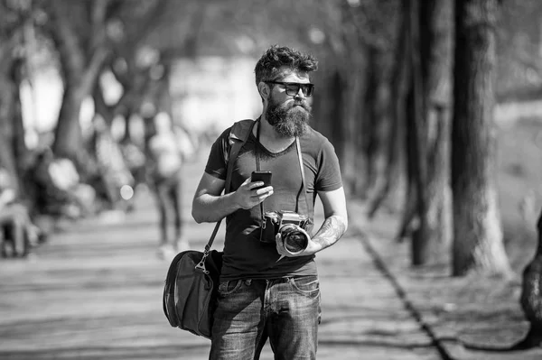 Young male photographer stroll around city in search for perfect picture. Bearded man in stylish eyewear holding vintage film camera and top-notch smartphone, modern gadgets vs old-fashioned devices