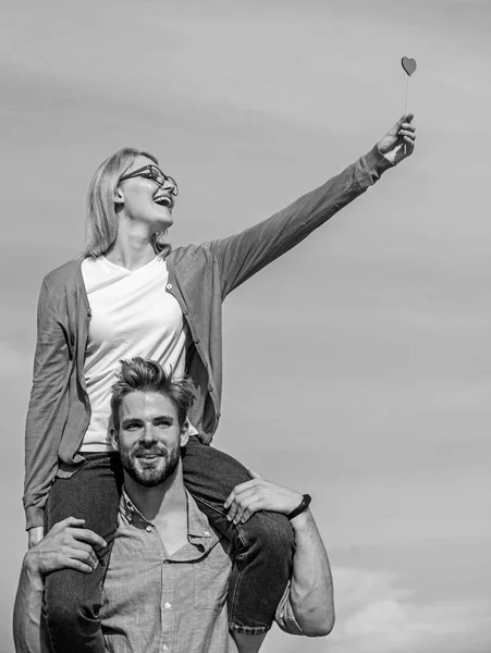 Homem carrega namorada nos ombros, fundo do céu. Casal feliz encontro se divertindo juntos. A mulher segura o coração no símbolo de pau do amor. Casal apaixonado andando ao ar livre dia ensolarado. Conceito de data romântica — Fotografia de Stock