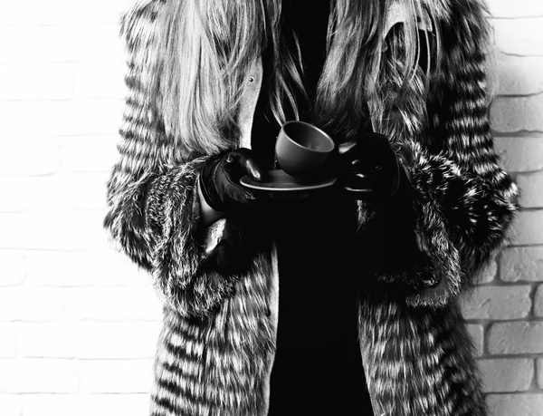 Γυναικεία χέρια της μόδας πλούσια γυναίκα ή κορίτσι με όμορφα μακριά ξανθά μαλλιά στη μέση παλτό από γκρι γούνα με μαύρα δερμάτινα γάντια κρατώντας καφέ κεραμικό φλιτζάνι καφέ σε φόντο στούντιο τούβλο — Φωτογραφία Αρχείου