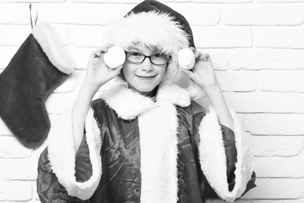 Młodych cute chłopiec santa claus w okularach w czerwony kapelusz sweter i nowy rok z ozdobny obsady Boże Narodzenie lub xmas lub rozruchu gospodarstwa marshmallows w pobliżu głowy na tle ściany Cegła biała — Zdjęcie stockowe