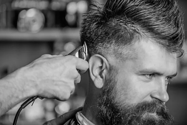 Barber works with hair clipper. Barbershop concept. Hands of barber with hair clipper, close up. Hipster bearded client getting hairstyle. Man with beard in hairdressers chair, dark background