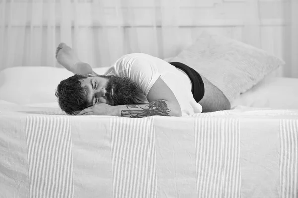 Tipo en la cara tranquila durmiendo en el borde de la cama en sábanas blancas. Hombre acostado en la cama, cortinas blancas en el fondo. Concepto siesta y siesta. Macho con barba y bigote durmiendo, relajándose, durmiendo la siesta, descansando — Foto de Stock