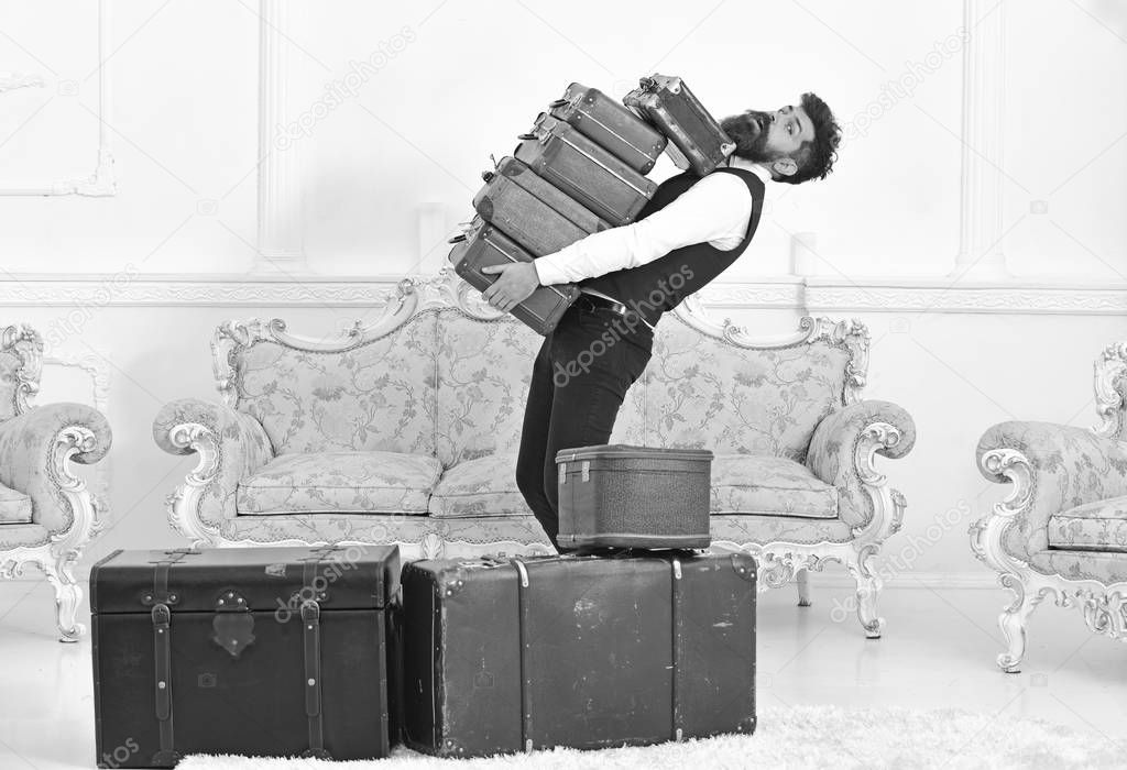 Man with beard and mustache wearing classic suit delivers luggage, luxury white interior background. Macho, elegant porter carries heavy pile of vintage suitcases. Butler and service concept