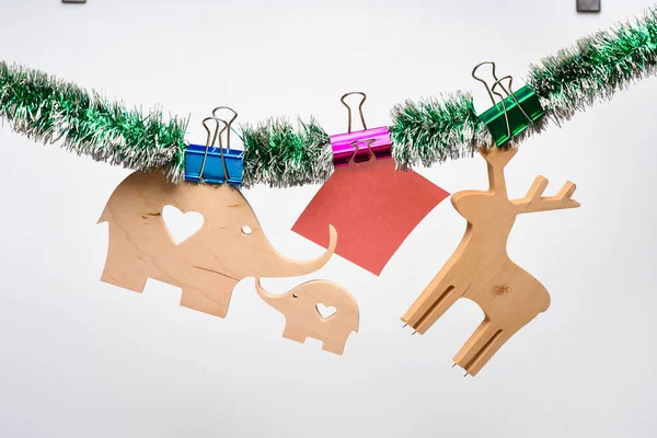 Concepto de decoración navideña. Decoraciones para las vacaciones de Navidad hechas de materiales naturales y oropel. Ciervo y elefante de madera con papel para nota cuelgan en oropel sobre fondo blanco —  Fotos de Stock