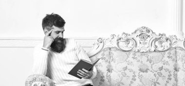 Sakal ve bıyık oturur barok tarzı kanepe, tutar kitap, beyaz duvar arka plan üzerinde olan adam. Maço yüz kitap okuma konusunda tutkulu gülümseyen üzerinde. Okuyucu kavramı. Adam zevk ile eski kitap okuma