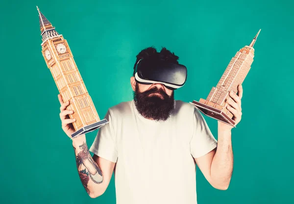Man on confused face study architecture or design in virtual reality. Guy in VR glasses holds Big Ben and Empire State Building. 3D design concept. Man with beard in VR glasses, green background