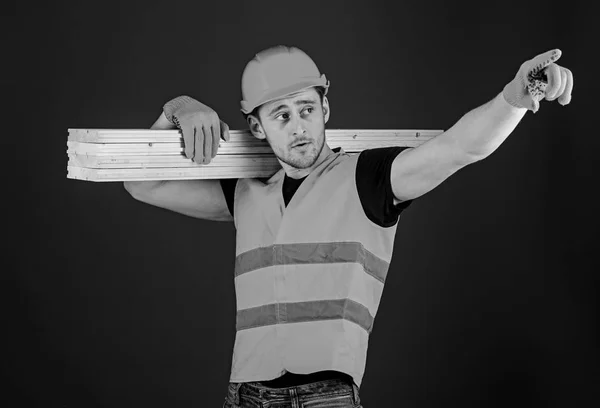 Houten materialen-concept. Timmerman, schrijnwerker, sterke bouwer op drukke gezicht draagt houten balk op de schouder. Man in de helm, helm en beschermende handschoenen wijzen richting, blauwe achtergrond — Stockfoto