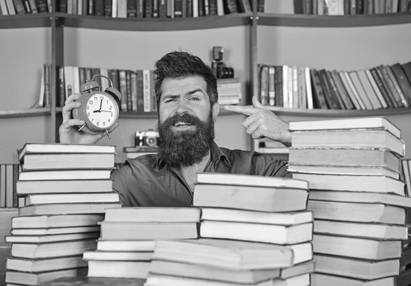 Meu, cientista a espreitar de montes de livros com despertador. Professor ou estudante com barba estudando na biblioteca. Homem na cara feliz apontando para o relógio, estantes de livros no fundo. Conceito de fluxo de tempo — Fotografia de Stock