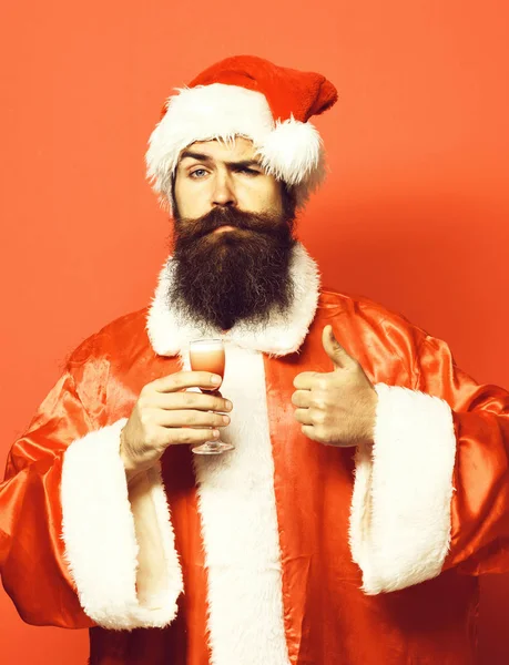 Hezký vousatý muž santa claus s dlouhým vousem na vážnou tváří drží sklo alkoholické střely v vánoční nebo svetr vánoční a novoroční klobouk znázorňující cool na červené studio pozadí — Stock fotografie