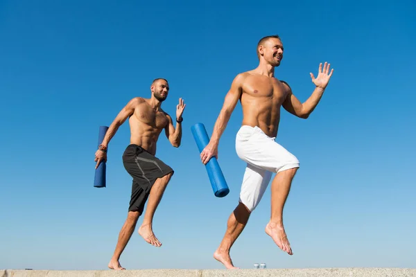 Adelante a un estilo de vida saludable. Hombres con esterilla de yoga capturados en movimiento fondo cielo azul. Deportista con estera corriendo. Ejecutar entrenamiento al aire libre. Los corredores se apresuran a estirar los músculos después del entrenamiento. Clases de yoga — Foto de Stock