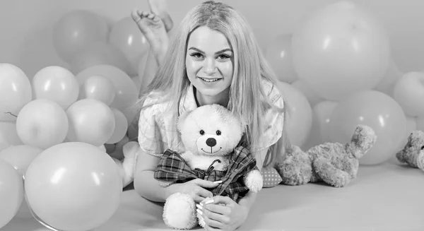 Blonde on smiling face relaxing with teddy bear toy. Woman cute celebrate birthday with balloons. Girl in pajama, domestic clothes lay near air balloons, pink background. Birthday girl concept