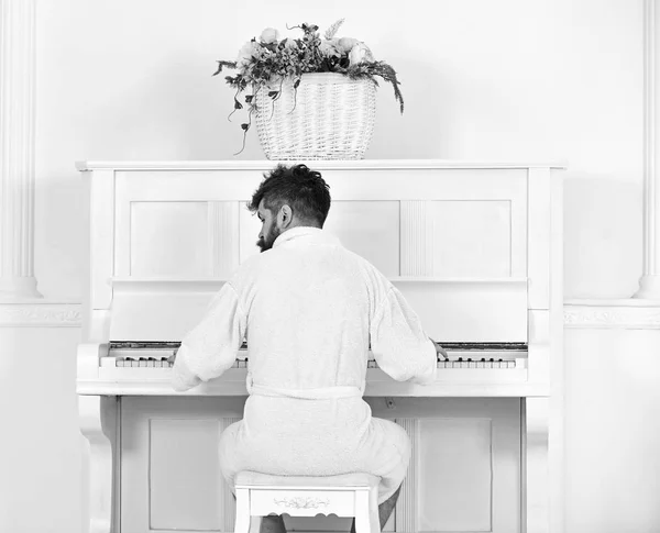 Man slaperig in de badjas zitten voor piano muziekinstrument in witte interieur op achtergrond, achteraanzicht. Getalenteerde muzikant concept. Man in badjas geniet ochtend tijdens het piano spelen — Stockfoto