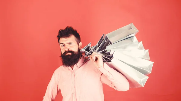 Hipster on sctrict face is shopping addicted or shopaholic. Guy shopping on sales season with discounts. Man with beard and mustache carries shopping bags on shoulder, red background. Shopping concept — Stock Photo, Image