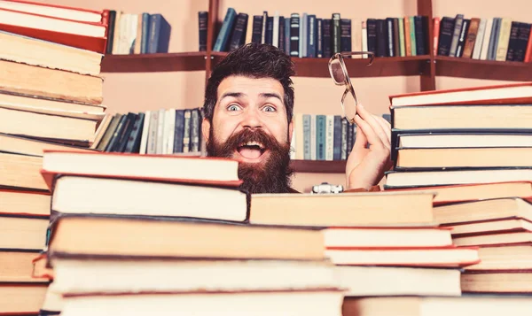 Homem no rosto feliz entre pilhas de livros na biblioteca, estantes de livros no fundo. Conceito de investigação científica. Professor ou estudante com barba usa óculos, senta-se à mesa com livros, desfocado — Fotografia de Stock