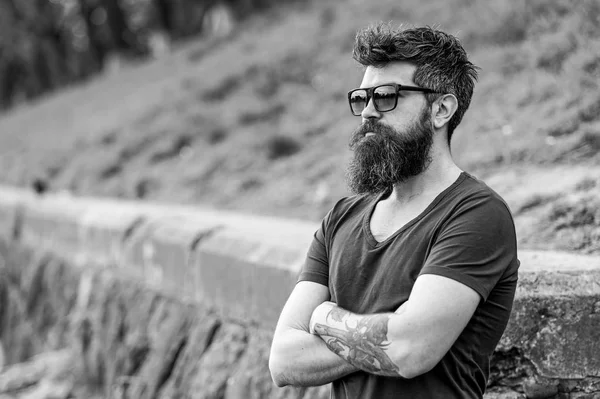 Barbearia e conceito de estilo. Homem com barba e bigode no rosto estrito, fundo da natureza, desfocado. Hipster com barba parece confiante enquanto está de pé ao ar livre. Homem barbudo usa óculos de sol modernos — Fotografia de Stock